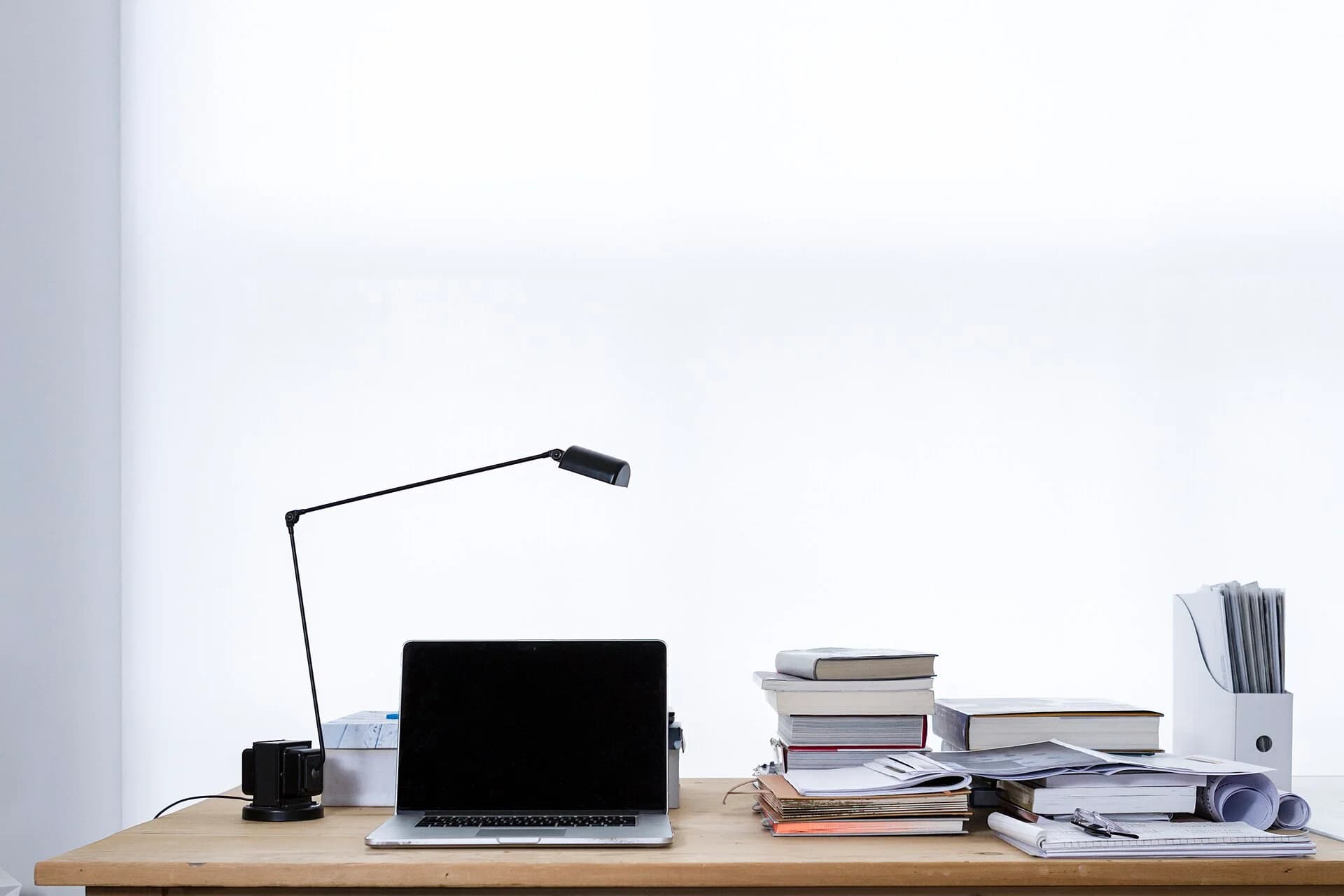 A desk lamp, computer and stacks of books by a bright window